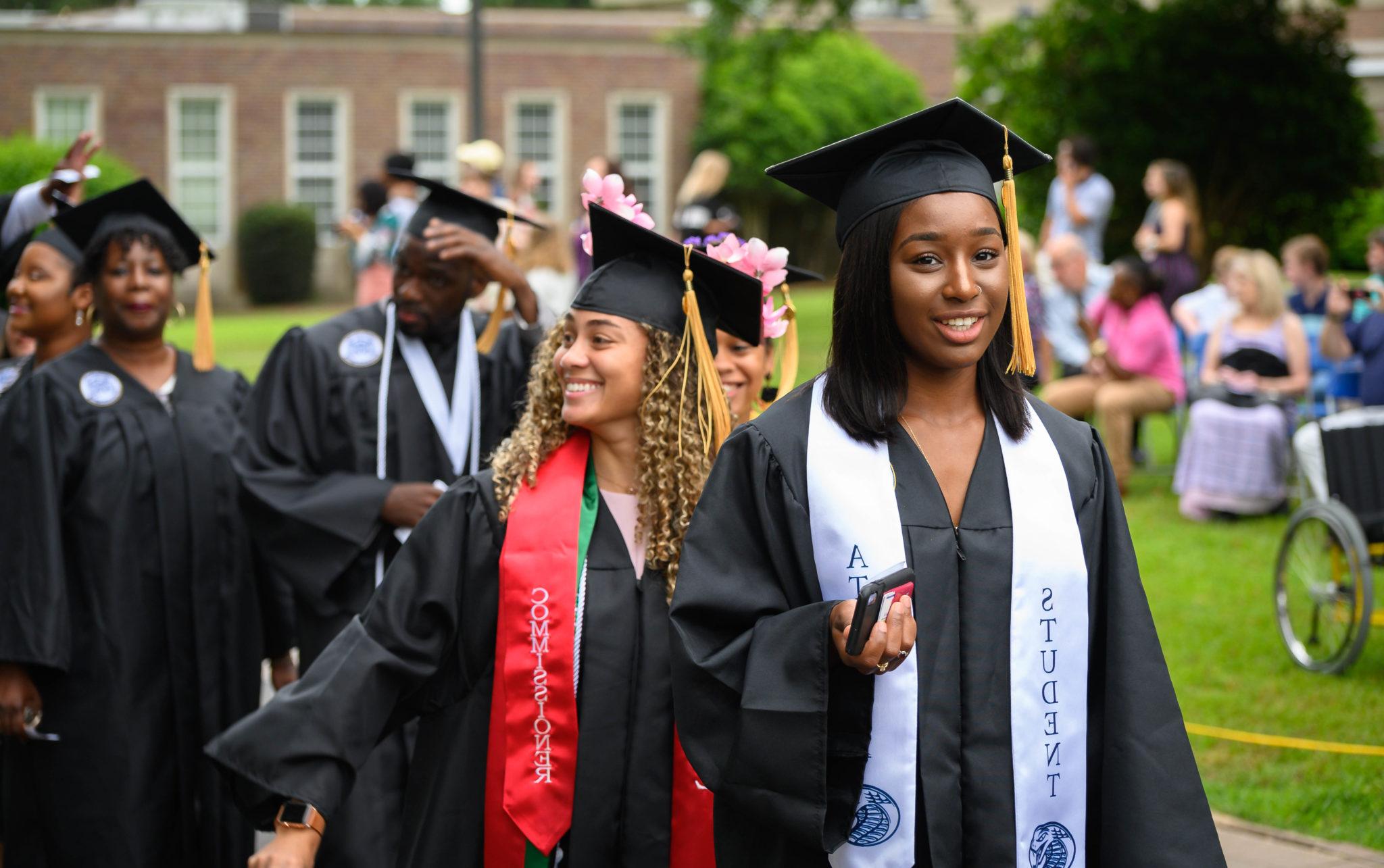 A student processes during an outdoor graduation ceremony, wearing a cap and gown